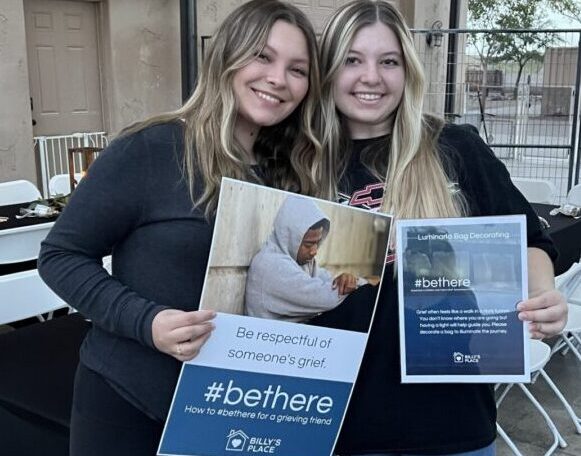 Two young women smiling at the camera while holding informational posters about supporting people in grief.