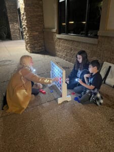 Three children play connect four at night, illuminated by a flashlight, outdoors near a stone building.