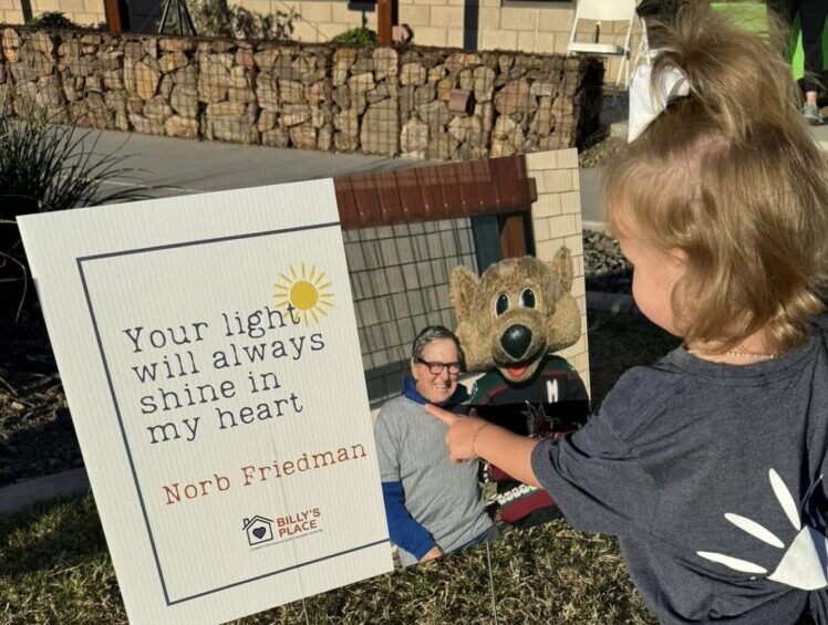 A young girl points at a memorial sign featuring a photo of a man with a mascot, inscribed with a tribute "your light will always shine in my heart, nord friedman.