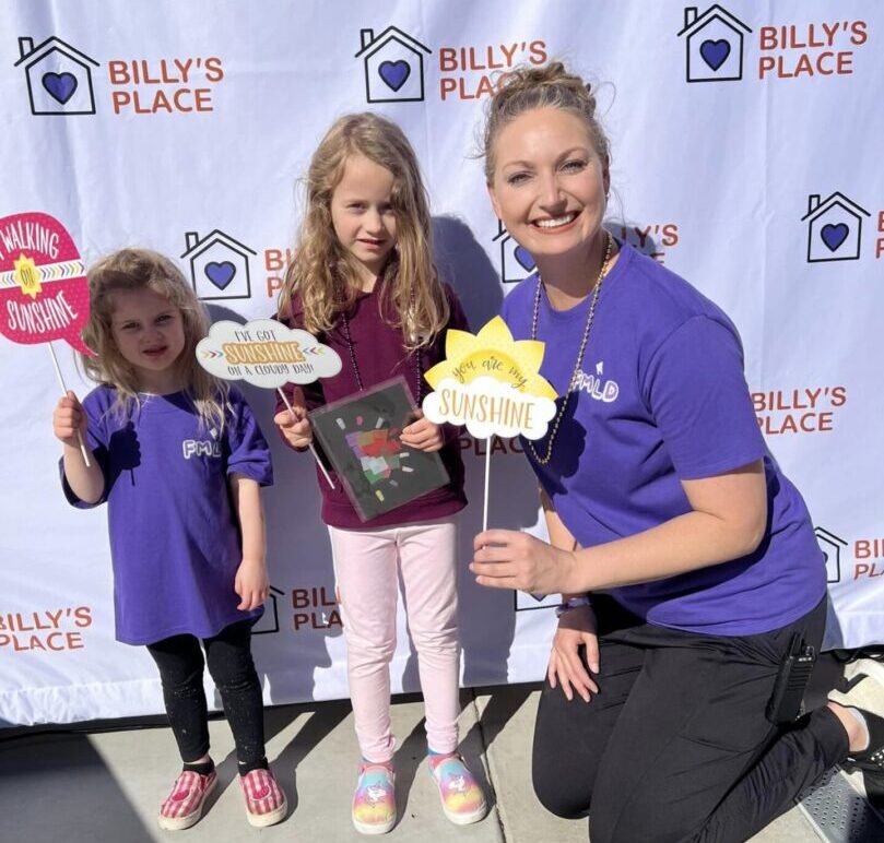 Two children and a woman holding "walking on sunshine" signs at an event with a backdrop labeled "billy's place.