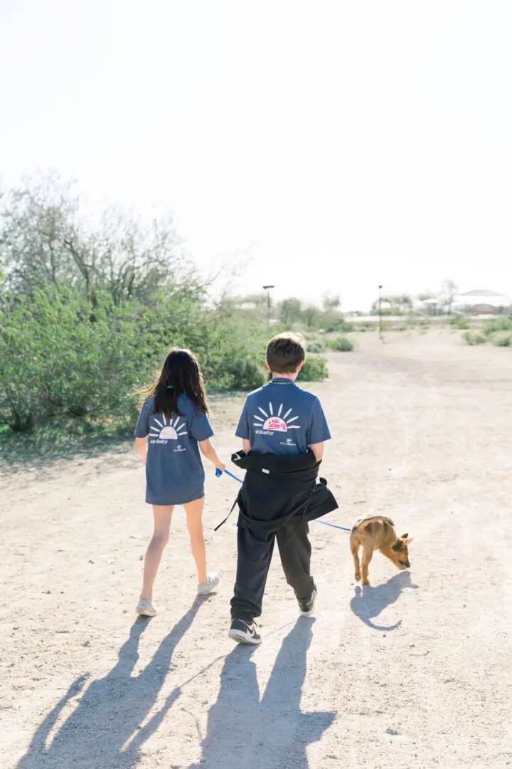 Two people walking a dog on a sandy path, wearing matching t-shirts and carrying a backpack.