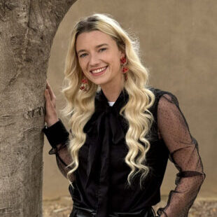 A smiling woman with blonde curly hair, wearing a black blouse and red earrings, posing by a tree.