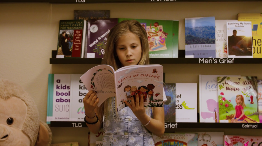 A young girl reads a book titled "death of a cupcake" in a bookstore, surrounded by various children's books on shelves.