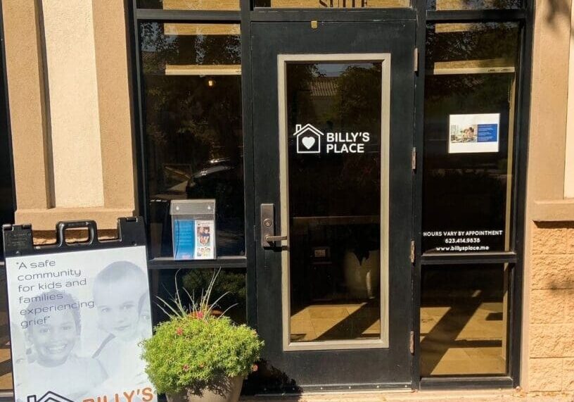 Glass door entrance to "billy's place" with a sidewalk sign featuring text and images, surrounded by a potted plant under a sunny sky.