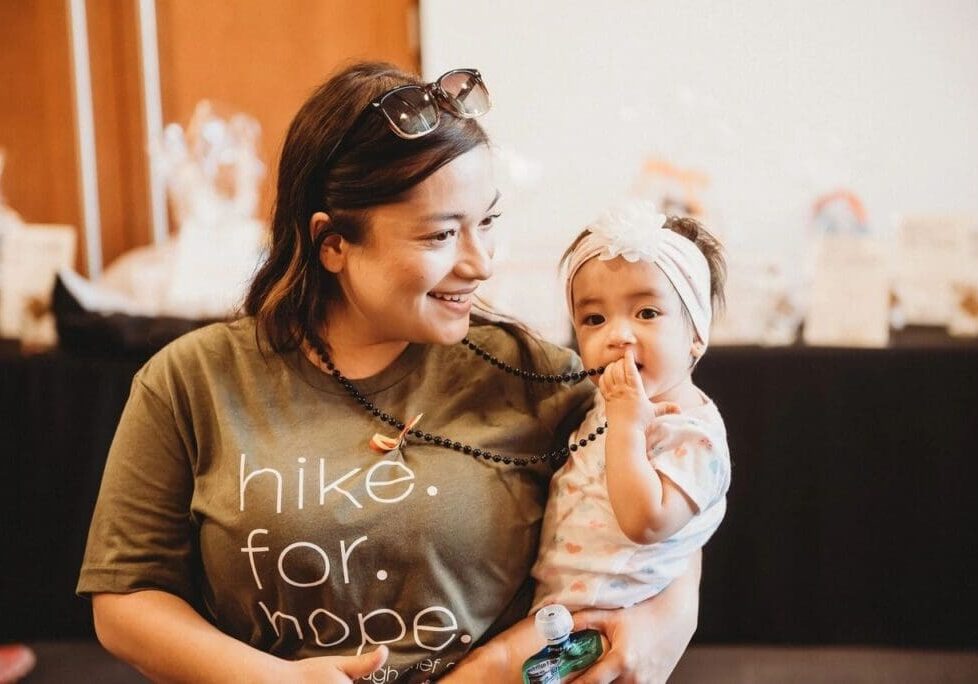 A woman holding a baby at an indoor event, both looking content; the woman wears a "hike for hope" t-shirt and the baby wears a headband.