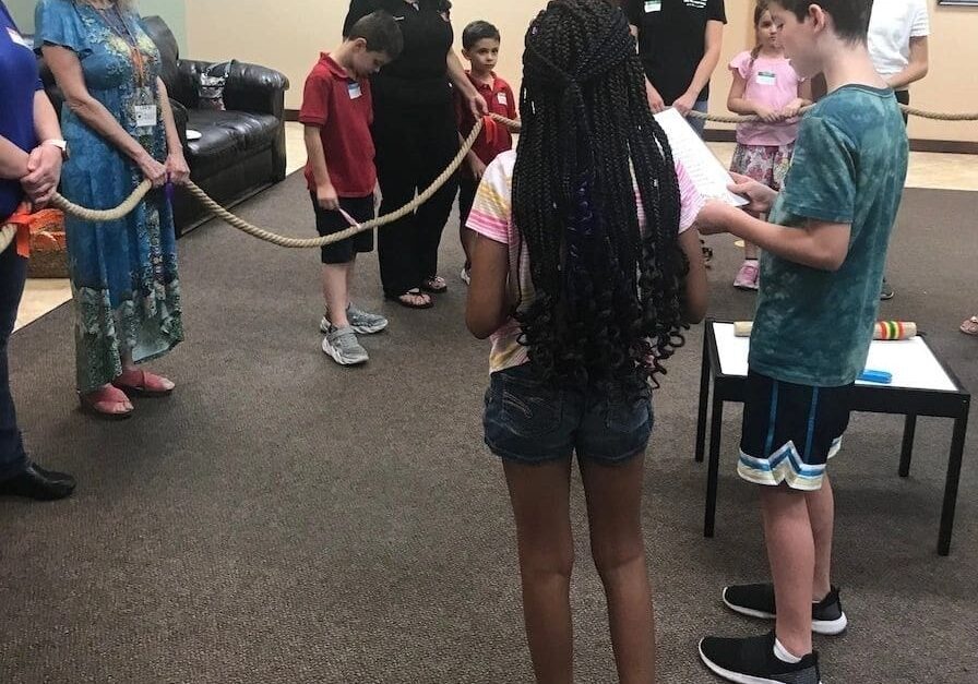 A group of children and adults in a room participating in a rope activity, focusing and collaborating together.