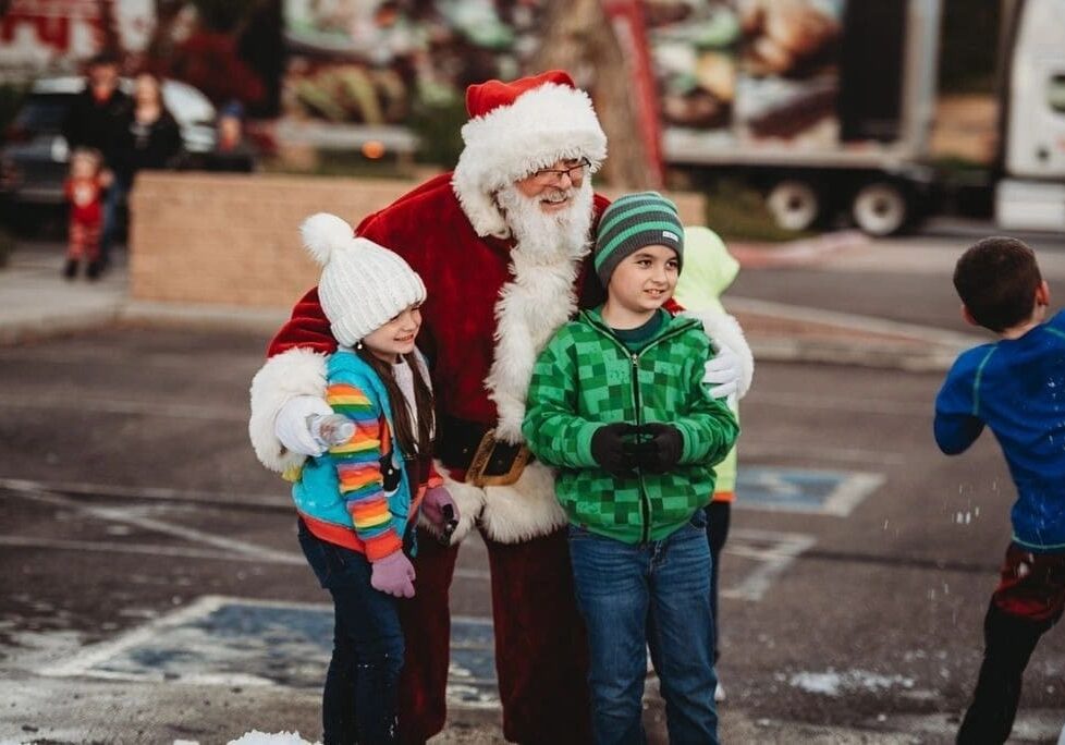 Santa claus embracing two children in winter clothing at an outdoor holiday event, with blurred festive background.