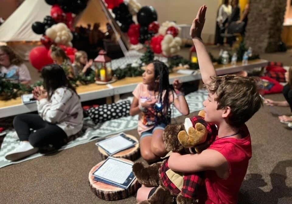 A child with a teddy bear raises their hand eagerly during a group indoor activity, surrounded by other children and festive red and black balloons.