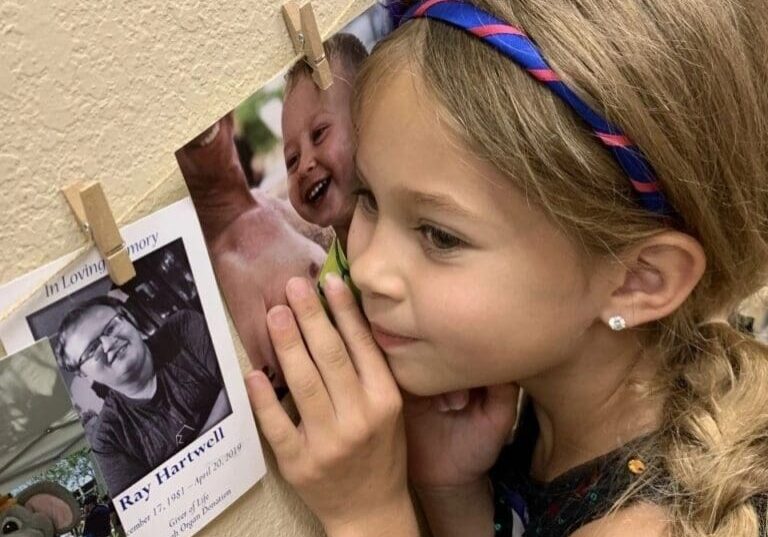 Young girl with a headband looking at photos pinned to a wall, touching a picture of a smiling man affectionately.