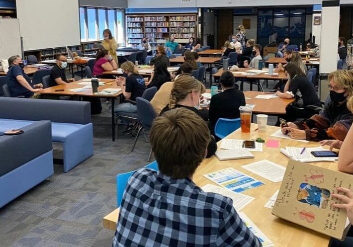 High school students in a library workshop, some seated at tables in groups, others on couches, with a teacher presenting at the front.