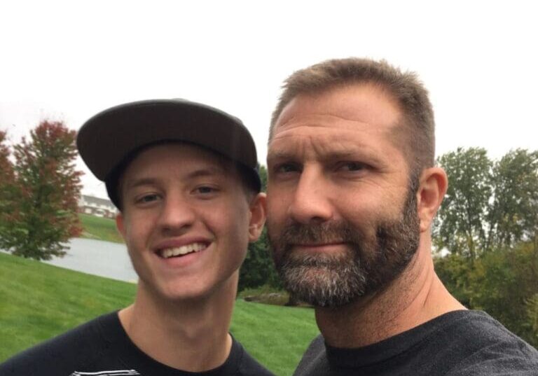Two men, one younger with a cap and one older with a beard, smiling and taking a selfie outdoors on a cloudy day.