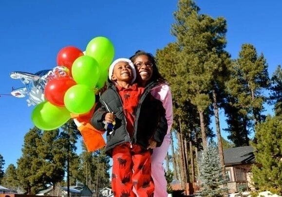 Two children smiling joyfully, holding colorful balloons outdoors on a sunny day with snow on the ground and pine trees in the background.