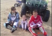 Three children in outdoor clothing sitting on sandy ground with a green atv in the background.